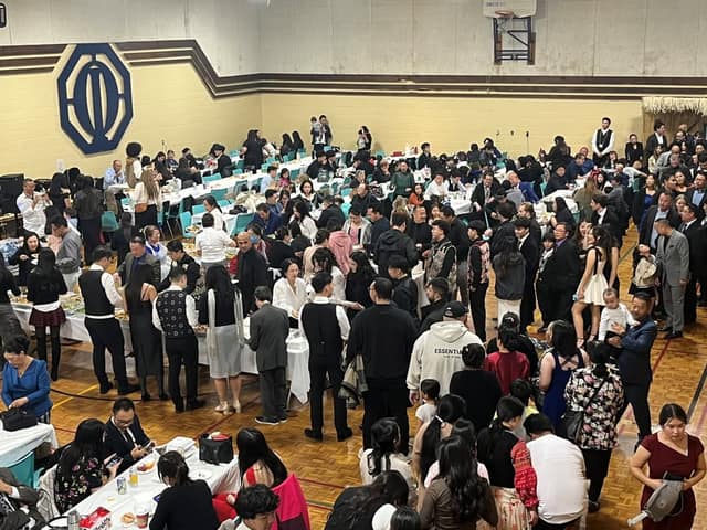 A crowded gymnasium full of people in dark and formal clothing, standing and seated at long tables during a buffet meal.