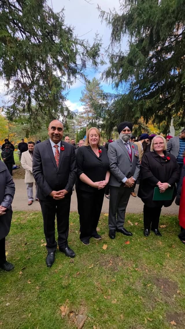 Two men and two women wearing poppies stand together on cemetery grass, with other attendees and tall evergreen trees behind them.