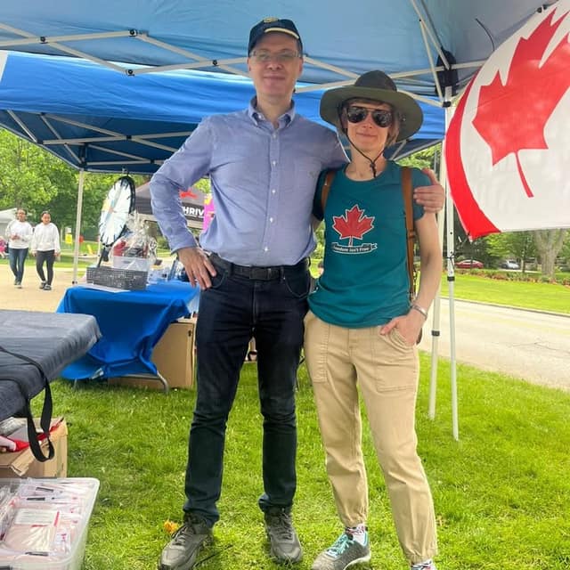 A tall man in a blue shirt and a woman in a hat and green maple leaf T-shirt stand together under a tent beside a Canadian flag.