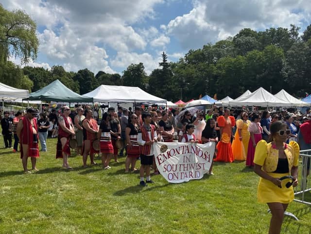 A crowd gathers on a park lawn as people in red traditional dress hold a banner reading Emontanosa in Southwest Ontario.