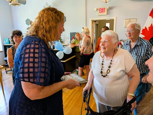 A woman in a navy dress holds a book and talks with an older woman using a walker as guests gather at the open house.