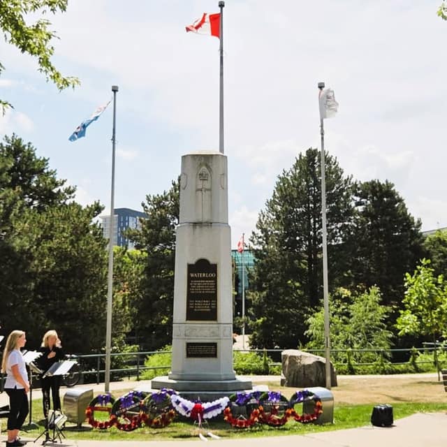 The Waterloo memorial stands among flagpoles on a sunny day, with wreaths at its base and two musicians with music stands at left.