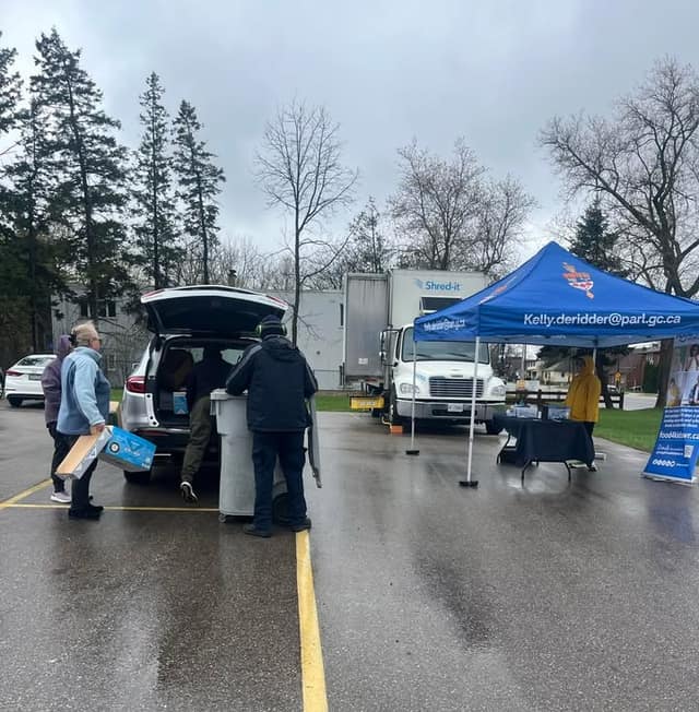 A resident carries a box toward a open SUV trunk beside a shred bin, with a Shred-it truck and blue tent in a wet parking lot.