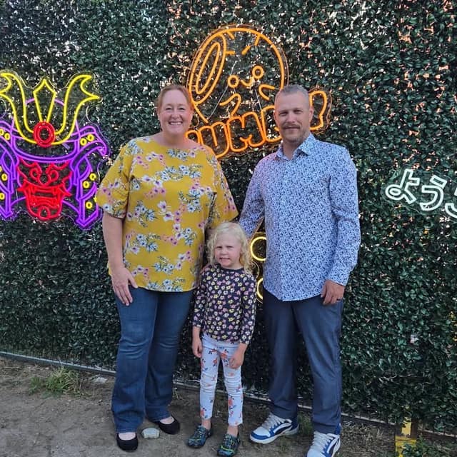 A woman in a yellow floral top, a man in a blue patterned shirt and a young child stand before a green hedge wall with neon signs.