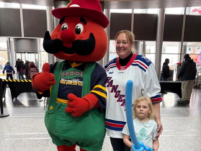 A woman in a Rangers jersey poses with a red Oktoberfest mascot in green overalls and a young girl holding a blue balloon sword.