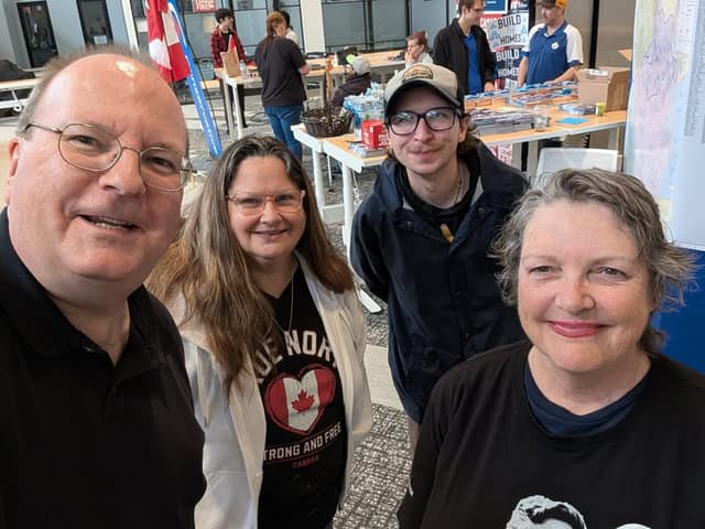 Four smiling people take a selfie in a campaign office with tables of materials and volunteers behind them.