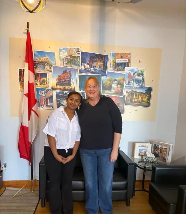 Setna, a co-op student, stands smiling beside a woman in a navy top and jeans in an office with a Canadian flag and a postcard mural.