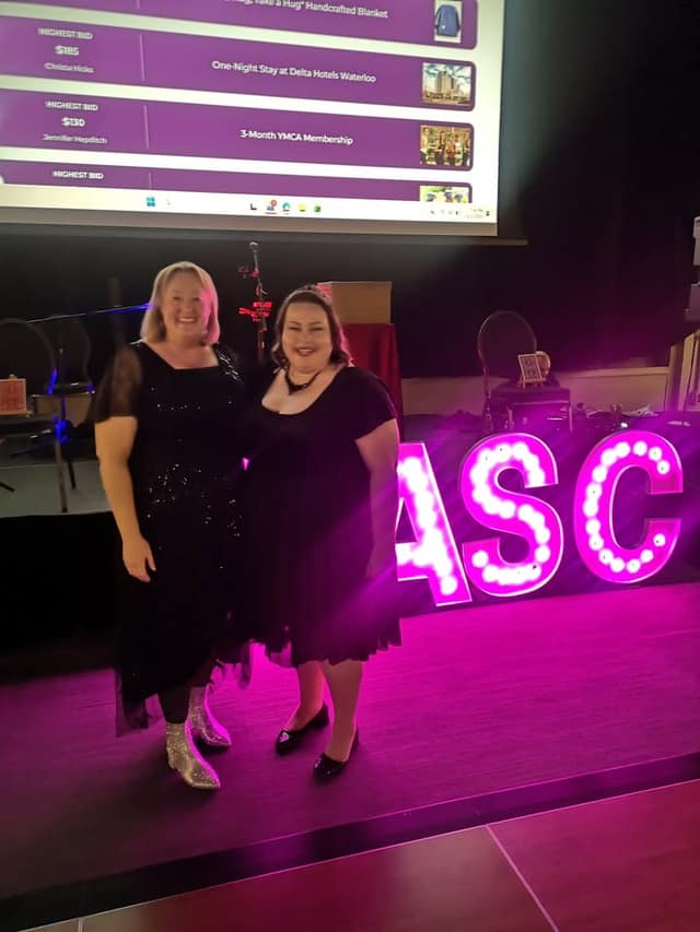 Two women in black dresses pose in front of glowing pink SASC marquee letters and a screen listing auction items and highest bids.