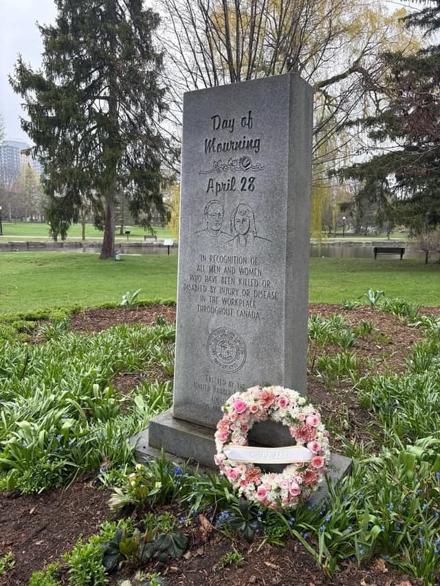 Granite Day of Mourning April 28 monument in a park, with a pink and white floral wreath at its base and spring plants around it.