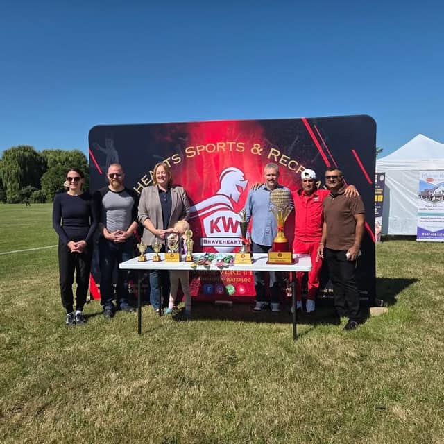 Six adults and a child stand behind a table of trophies and medals in front of a KW Bravehearts banner on a sunny field.