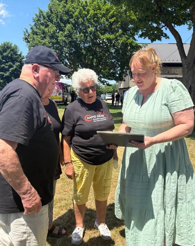 A woman in a green dress holds a framed certificate while two people in black Rapid Gear "Proudly Canadian" shirts look at it.