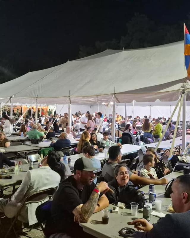 A crowd sits at long tables under a large white tent at night, with an Armenian flag at the right edge and drinks on the tables.