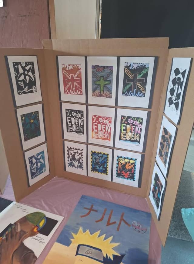 A cardboard display of block prints, including flowers, crosses, "Eden" lettering and 50c stamps, with a painting of an anime character in front.