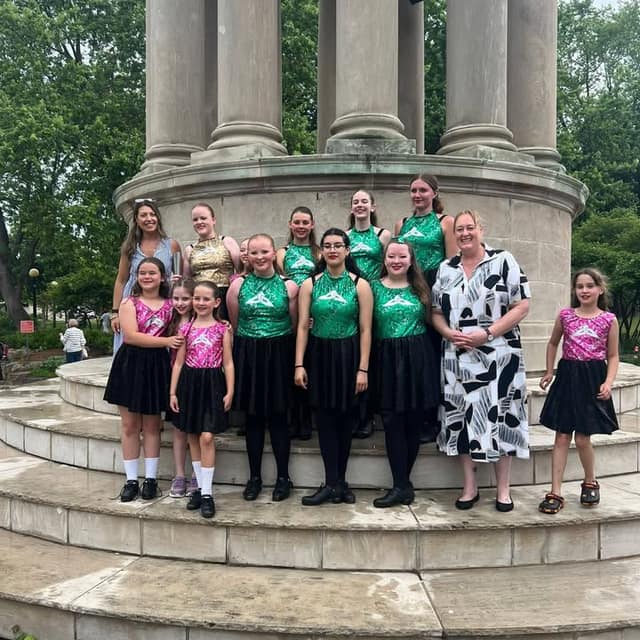 A group of girls in green or pink sparkly tops and black skirts pose with two women on the round stone steps of a pillared monument.