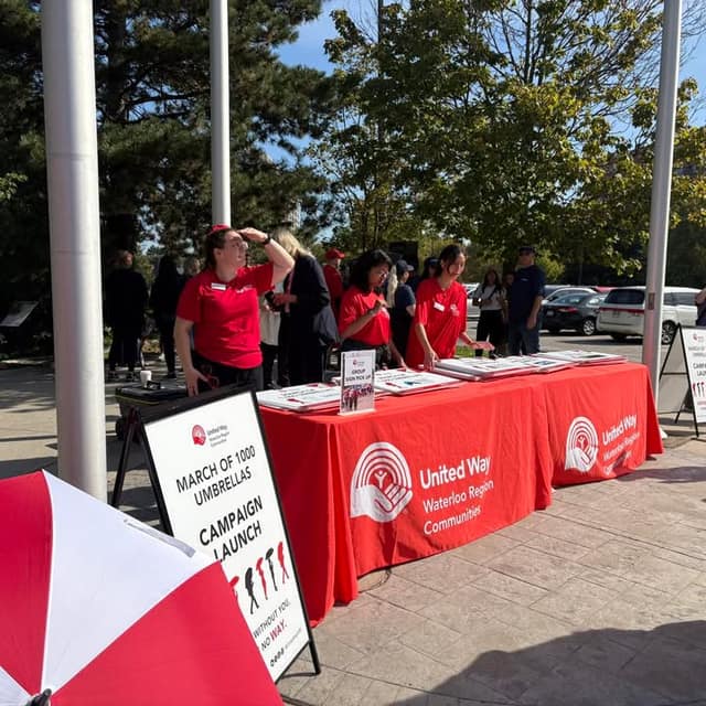 Volunteers in red shirts stand behind a red United Way Waterloo Region Communities table with a sign reading "March of 1000 Umbrellas Campaign Launch".