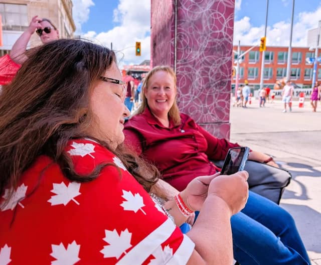 A smiling woman in a dark red shirt sits beside a woman in a maple leaf top who looks at a phone, with a crowd behind.