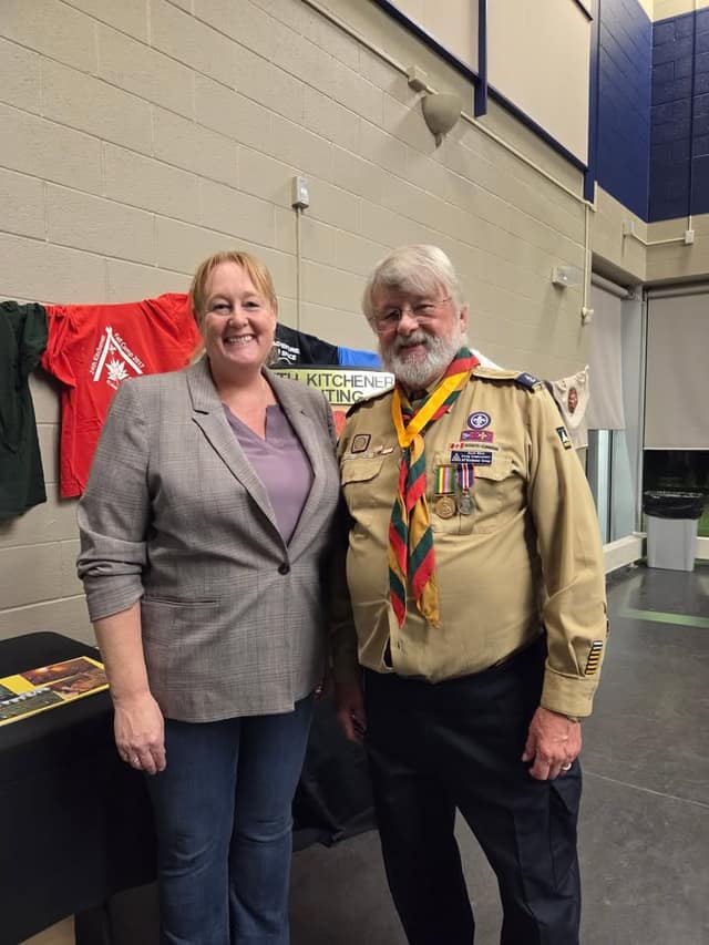 A smiling woman in a gray blazer stands beside an older bearded Scout leader in a tan uniform with a neckerchief and medals, in front of a display table.
