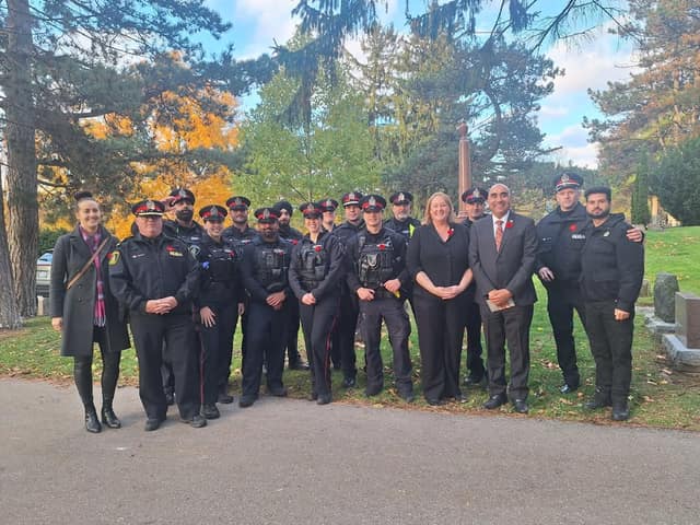 A woman and a man in a suit pose with about a dozen uniformed police officers and a civilian on a cemetery lawn in autumn.