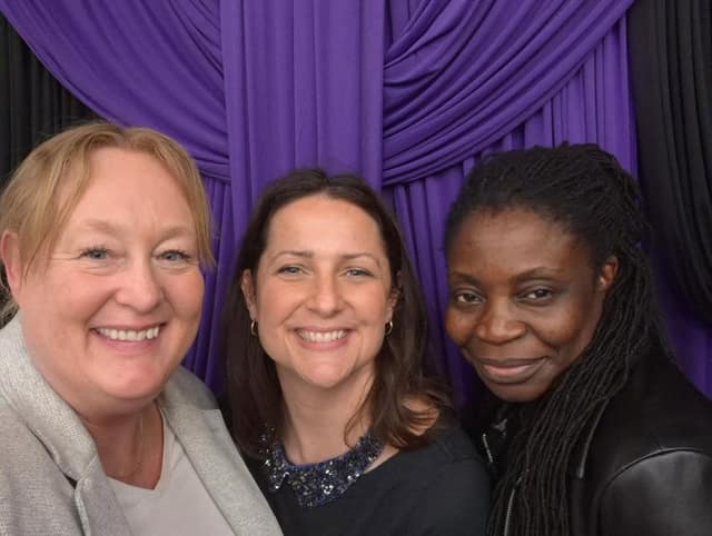 Three smiling women pose close together for a selfie in front of purple and black draped curtains.