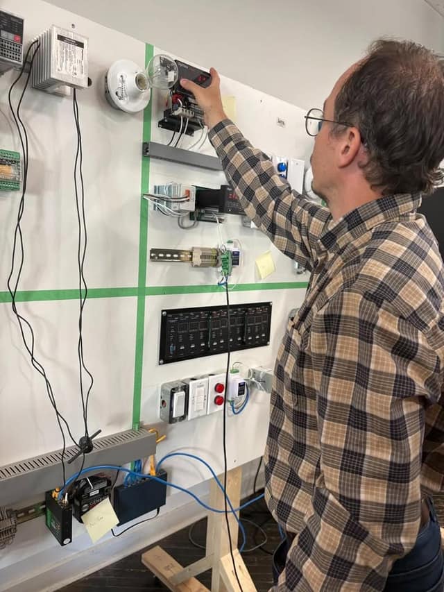 A man in a plaid shirt reaches up to adjust a device on a wall-mounted board of controllers, switches, light bulbs and cables.