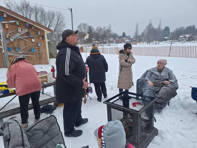 Residents stand and sit around a mesh fire pit in the snow, one roasting a marshmallow, beside a shed covered in birdhouses.