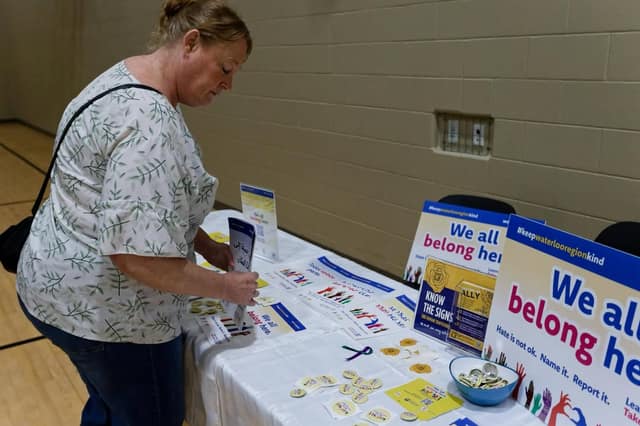 A woman in a leaf-patterned blouse picks up a brochure from a table with "We all belong here" signs, buttons and flyers.
