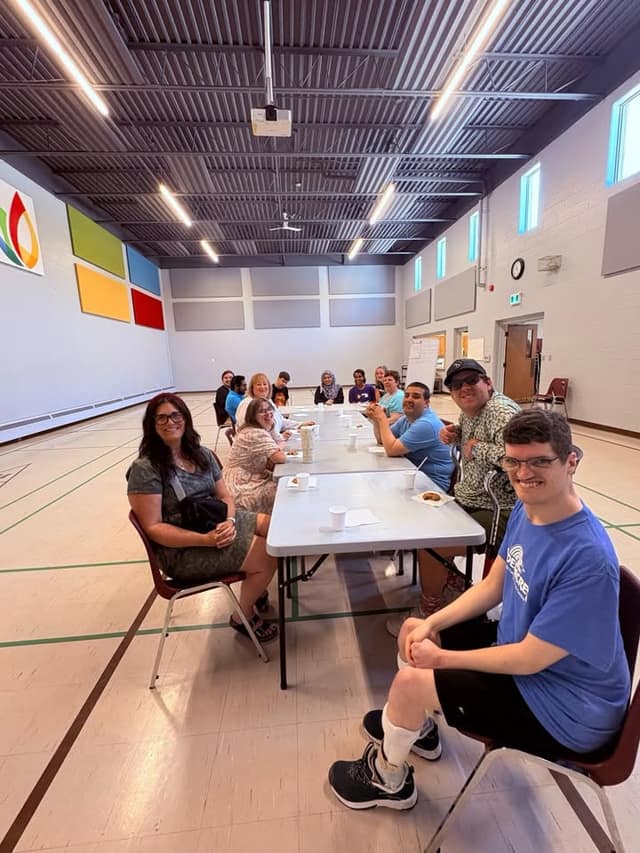 Smiling participants and staff sit along a long folding table in a bright gymnasium, looking toward the camera.