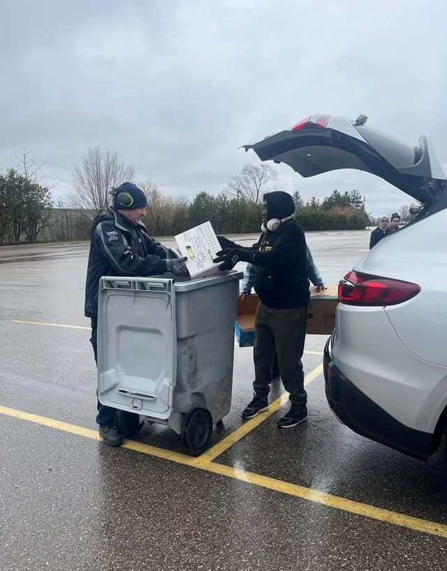 Two volunteers in rain gear lift boxes of paper into a grey shred bin beside an open SUV in a wet parking lot.
