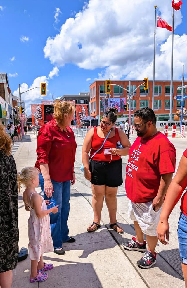 A woman in a red blouse talks with a woman and a man in red shirts while a small girl looks on, on a sunny street.