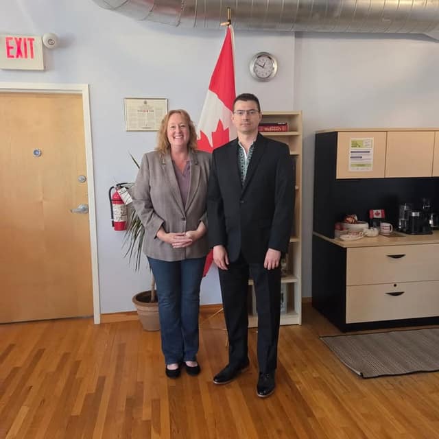 Kelly DeRidder and a man in a dark suit stand smiling in front of a Canadian flag in an office with a wood floor.