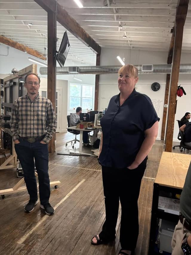 A woman in a navy blouse and a man in a plaid shirt stand in an open office with exposed wood beams while staff work at desks behind.