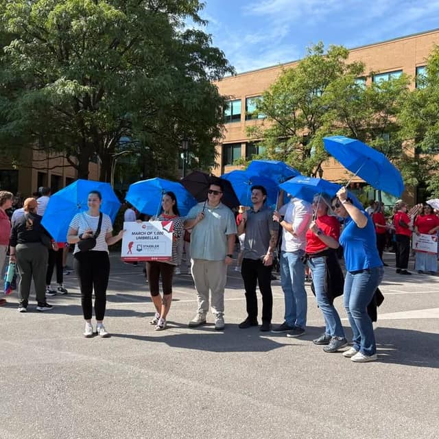 Participants in blue shirts hold blue umbrellas outdoors, lined up beside a sign reading "March of 1,000 Umbrellas" near a brick building.