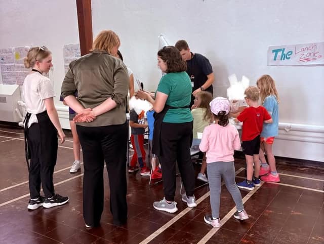 A woman in an olive shirt and camp staff stand around a table with several children doing an activity in a gym, beside a Lunch Zone sign.