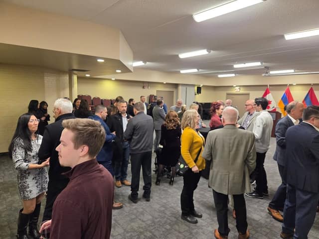 Guests stand and chat in small groups in a carpeted hall, with Canadian and Armenian flags and a podium visible in the background.