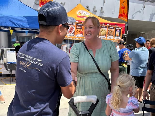 A woman in a green dress smiles and shakes hands with a man in a navy shirt while a girl stands nearby at a food tent.
