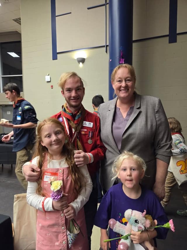 A smiling woman in a gray blazer stands with a young Scout leader in a red uniform and two girls holding flowers, one with a stuffed toy.