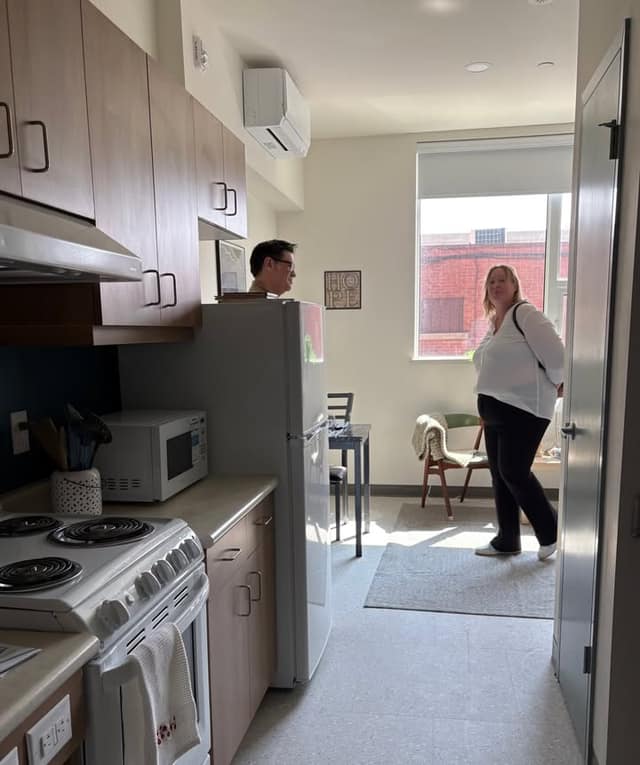 A small apartment kitchen with wood cabinets, a white fridge and stove; a woman stands by a sunny window while a man looks on from behind the fridge.