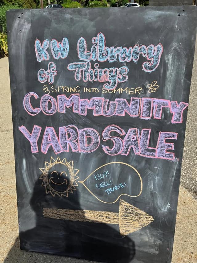 Close view of a chalkboard sign reading KW Library of Things, Spring into Summer Community Yard Sale, with a sun and Buy! Sell! Trade!