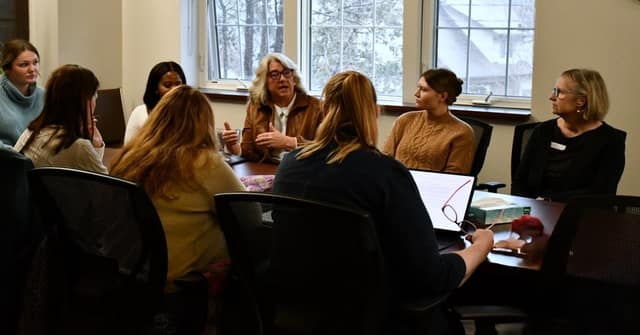 Seven women sit around a table in a bright room with windows as one woman in a tan jacket speaks and gestures to the group.