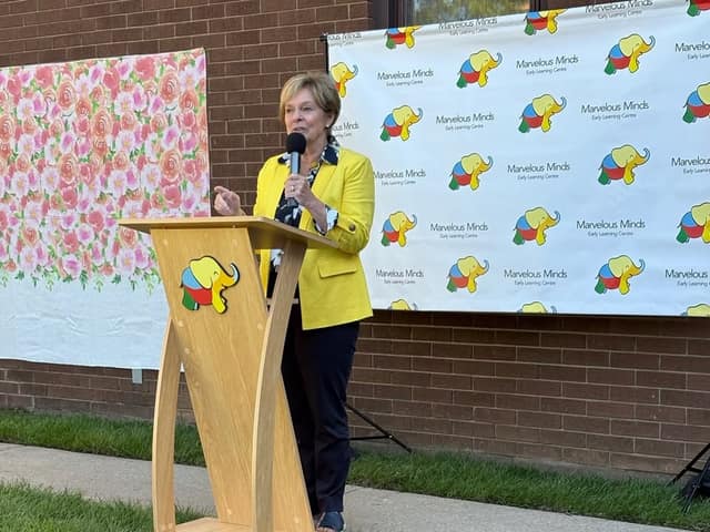 A woman in a yellow blazer speaks into a microphone at a wooden podium in front of a Marvelous Minds Early Learning Centre banner.