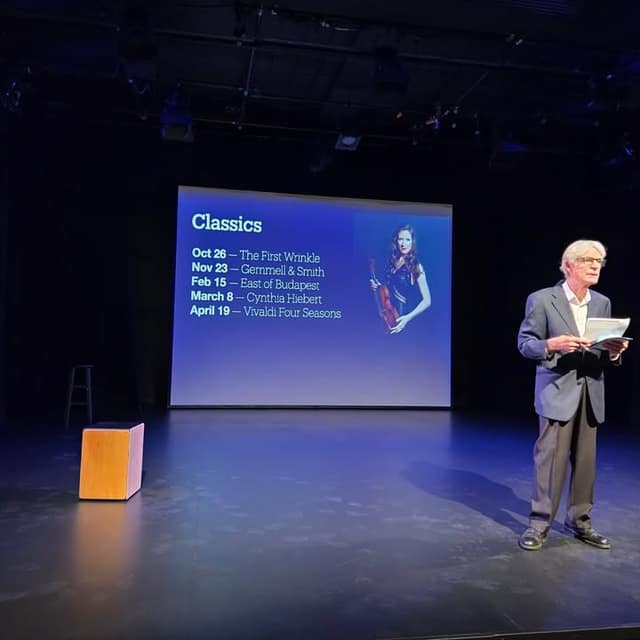 A man in a gray blazer holds papers on a dark stage beside a screen titled Classics listing concerts and a violinist photo.