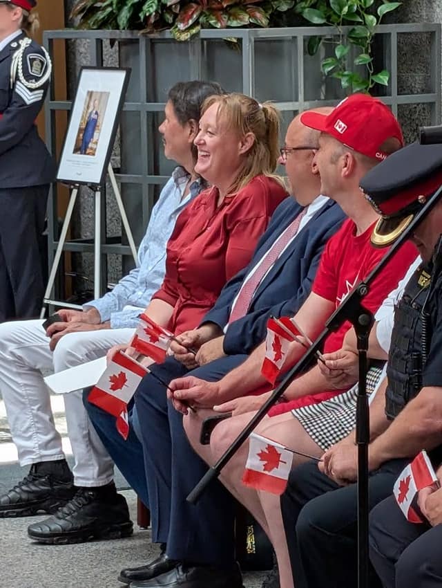 People seated in a row hold small Canadian flags and laugh, with a framed portrait on an easel and an officer behind them.