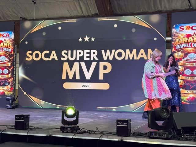 A woman in a colourful tunic holds papers beside a host with a microphone on stage, before a screen reading SOCA Super Woman MVP 2026.