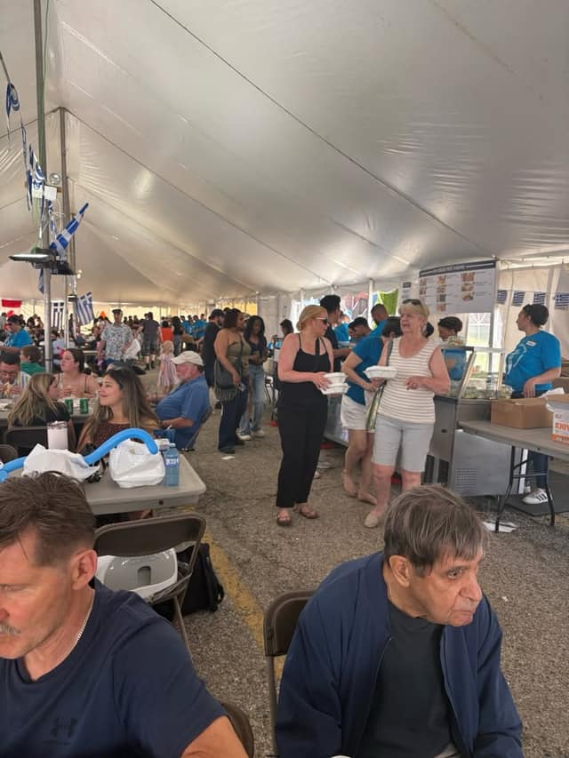 A crowded festival tent with people seated at tables and standing in line, Greek flags overhead and a menu board in the background.