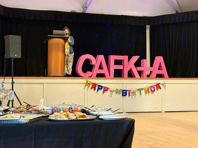 A person speaks at a podium on a stage beside large pink letters reading CAFK+A, with a Happy Birthday banner and a food table in front.