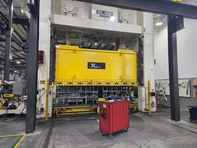 A huge Blow industrial press with a yellow XL Tool die section, and a red rolling tool cart on the shop floor in front.