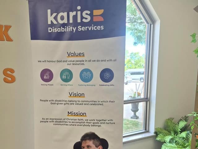 A Karis Disability Services banner listing its values, vision and mission, standing beside a window and a potted fern.