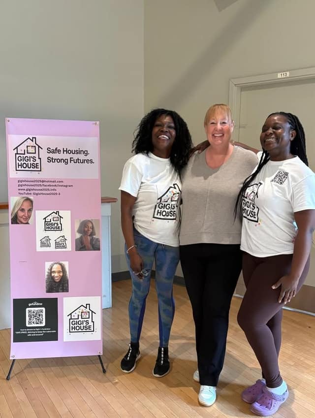 Three smiling women stand together beside a pink Gigi's House banner reading "Safe Housing. Strong Futures."; two wear Gigi's House T-shirts.