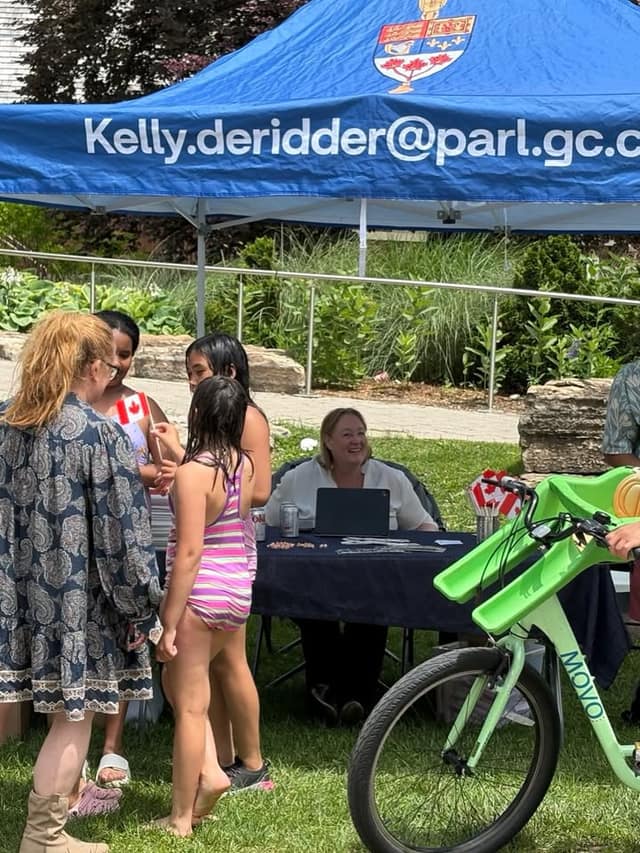Children in swimsuits and a woman gather at a table under a blue tent with an email address, while a woman sits behind it near a green bike.