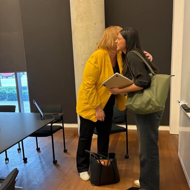 A woman in a yellow jacket hugs a smiling woman carrying a green tote bag and laptop in a meeting room with a black table.